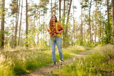 Parlak bir sırt çantasıyla neşeli bir kadın güneşli bir ormanda doğanın tadını çıkarıyor. Yürüyüş yolunda güzel bir bayan gezgin. Yürüyüş, dinlenme ve doğa konsepti.