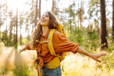 Parlak bir sırt çantasıyla neşeli bir kadın güneşli bir ormanda doğanın tadını çıkarıyor. Yürüyüş yolunda güzel bir bayan gezgin. Yürüyüş, dinlenme ve doğa konsepti.
