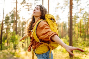 Parlak bir sırt çantasıyla neşeli bir kadın güneşli bir ormanda doğanın tadını çıkarıyor. Yürüyüş yolunda güzel bir bayan gezgin. Yürüyüş, dinlenme ve doğa konsepti.