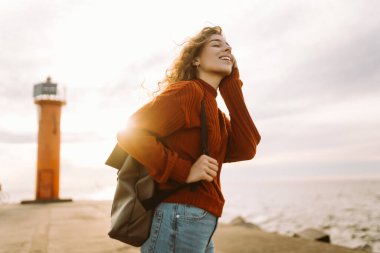 Kazaklı ve sırt çantalı sakin bir kadın rıhtımda deniz esintisinin ve temiz havada gün batımının tadını çıkarıyor. Güzel bir kadın doğada kendini özgür ve rahat hisseder. Doğa konsepti, rahatlama..