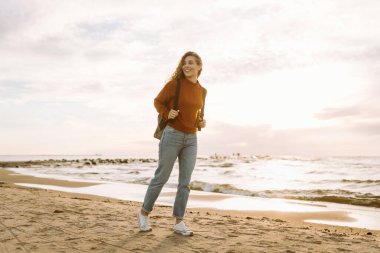 Kazaklı ve sırt çantalı sakin bir kadın rıhtımda deniz esintisinin ve temiz havada gün batımının tadını çıkarıyor. Güzel bir kadın doğada kendini özgür ve rahat hisseder. Doğa konsepti, rahatlama..