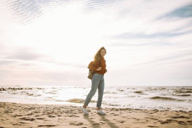 Kazaklı ve sırt çantalı sakin bir kadın rıhtımda deniz esintisinin ve temiz havada gün batımının tadını çıkarıyor. Güzel bir kadın doğada kendini özgür ve rahat hisseder. Doğa konsepti, rahatlama..