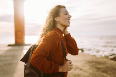 Kazaklı ve sırt çantalı sakin bir kadın rıhtımda deniz esintisinin ve temiz havada gün batımının tadını çıkarıyor. Güzel bir kadın doğada kendini özgür ve rahat hisseder. Doğa konsepti, rahatlama..