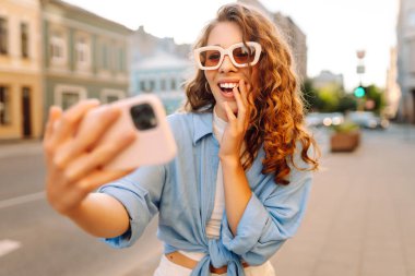 Güneş gözlüklü ve telefonlu genç bir kadın güneşli bir sokakta selfie çekiyor. Güzel kadın blogları ve gün batımında şehrin sokaklarında gezinerek eğleniyor. Yaşam biçimi.
