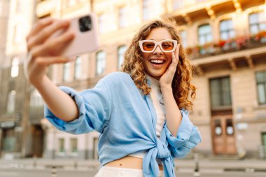 Güneş gözlüklü ve telefonlu genç bir kadın güneşli bir sokakta selfie çekiyor. Güzel kadın blogları ve gün batımında şehrin sokaklarında gezinerek eğleniyor. Yaşam biçimi.
