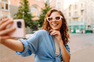 Güneş gözlüklü ve telefonlu genç bir kadın güneşli bir sokakta selfie çekiyor. Güzel kadın blogları ve gün batımında şehrin sokaklarında gezinerek eğleniyor. Yaşam biçimi.
