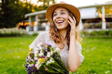 Şapkalı neşeli bir kadın portresi ve güneşli bir parkta yürüyen bir buket kır çiçeğiyle. Çiçekleri olan güzel bir turist gün batımının tadını çıkarıyor. Sürpriz kavramı, rahatlama, güzellik..
