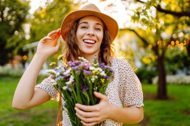 Şapkalı neşeli bir kadın portresi ve güneşli bir parkta yürüyen bir buket kır çiçeğiyle. Çiçekleri olan güzel bir turist gün batımının tadını çıkarıyor. Sürpriz kavramı, rahatlama, güzellik..