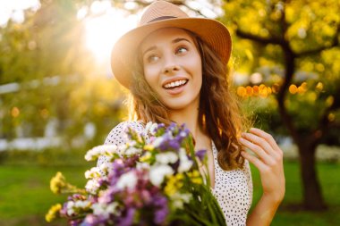 Şapkalı neşeli bir kadın portresi ve güneşli bir parkta yürüyen bir buket kır çiçeğiyle. Çiçekleri olan güzel bir turist gün batımının tadını çıkarıyor. Sürpriz kavramı, rahatlama, güzellik..