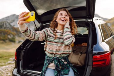 Araba bagajında telefonla gezen, selfie çeken ya da dağ manzarasına karşı blog yazan bir gezgin. Gün batımının keyfini çıkaran güzel bir kadın ve araba yolculuğunda doğa. Ulaşım, macera ve günlük kavramı.