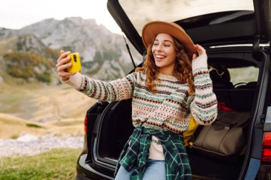 Araba bagajında telefonla gezen, selfie çeken ya da dağ manzarasına karşı blog yazan bir gezgin. Gün batımının keyfini çıkaran güzel bir kadın ve araba yolculuğunda doğa. Ulaşım, macera ve günlük kavramı.