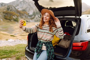 Araba bagajında telefonla gezen, selfie çeken ya da dağ manzarasına karşı blog yazan bir gezgin. Gün batımının keyfini çıkaran güzel bir kadın ve araba yolculuğunda doğa. Ulaşım, macera ve günlük kavramı.