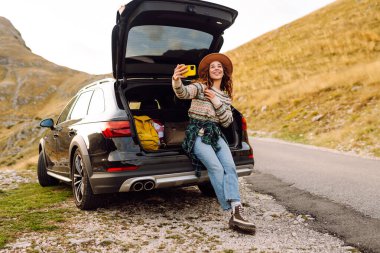 Araba bagajında telefonla gezen, selfie çeken ya da dağ manzarasına karşı blog yazan bir gezgin. Gün batımının keyfini çıkaran güzel bir kadın ve araba yolculuğunda doğa. Ulaşım, macera ve günlük kavramı.