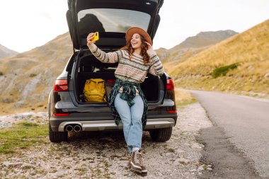 Araba bagajında telefonla gezen, selfie çeken ya da dağ manzarasına karşı blog yazan bir gezgin. Gün batımının keyfini çıkaran güzel bir kadın ve araba yolculuğunda doğa. Ulaşım, macera ve günlük kavramı.
