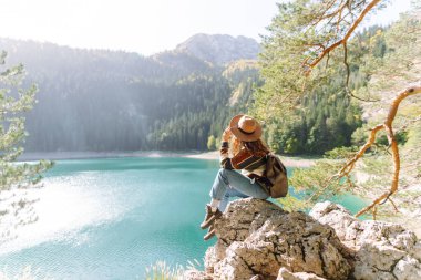 Şapkalı ve sırt çantalı bir kadın dağcı yüksek bir dağ gölüne bakan bir kayanın üzerinde oturuyor. Genç kadın özgürlüğü hissediyor ve dağ manzarasının tadını çıkarıyor. Yürüyüş ve doğa konsepti.