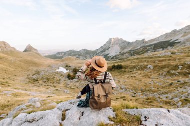 Sırt çantalı, şapkalı ve otantik kazaklı mutlu bayan yürüyüşçü dağ yolunda dağ manzarasının tadını çıkarıyor. Genç bayan yürüyüşçü inanılmaz manzaralarla çevrili özgürlüğü hissediyor. Etkin yaşam biçimi.