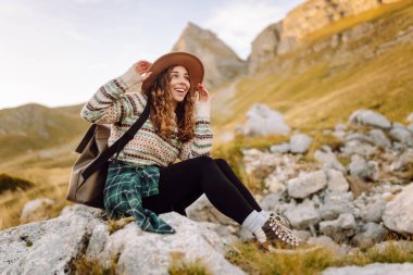 Sırt çantalı, şapkalı ve otantik kazaklı mutlu bayan yürüyüşçü dağ yolunda dağ manzarasının tadını çıkarıyor. Genç bayan yürüyüşçü inanılmaz manzaralarla çevrili özgürlüğü hissediyor. Etkin yaşam biçimi.