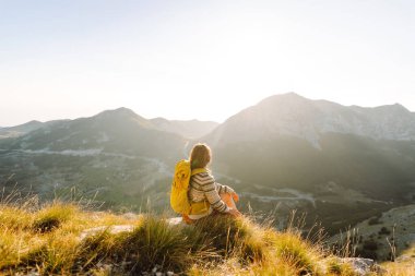 Parlak bir sırt çantası olan neşeli bir kadın güzel bir dağ yolunda yürüyor. Güzel bayan gezgin dağlarda günbatımının tadını çıkarıyor ve özgürlüğü hissediyor. Yürüyüş, özgürlük konsepti.