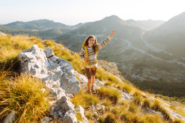 Parlak bir sırt çantası olan neşeli bir kadın güzel bir dağ yolunda yürüyor. Güzel bayan gezgin dağlarda günbatımının tadını çıkarıyor ve özgürlüğü hissediyor. Yürüyüş, özgürlük konsepti.