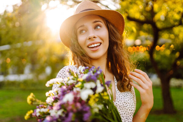 Портрет веселой женщины в шляпе с букетом диких цветов, гуляющей в солнечном парке. Турист с цветами, наслаждающийся закатом на свежем воздухе. Концепция неожиданности, расслабления, красоты.
