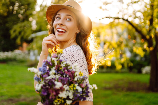 Портрет веселой женщины в шляпе с букетом диких цветов, гуляющей в солнечном парке. Турист с цветами, наслаждающийся закатом на свежем воздухе. Концепция неожиданности, расслабления, красоты.