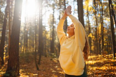 Günbatımında sonbahar ormanında parlak kazaklı genç kadın yoga yapıyor. Atletik kadın egzersiz yapıyor, dışarıda esniyor. Meditasyon, yoga ve rahatlama konsepti.