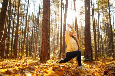 Günbatımında sonbahar ormanında parlak kazaklı genç kadın yoga yapıyor. Atletik kadın egzersiz yapıyor, dışarıda esniyor. Meditasyon, yoga ve rahatlama konsepti.