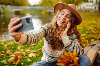 Şapkalı neşeli kadın, sonbahar parkında kırmızı bir battaniyenin üzerinde otururken telefonuyla selfie çekiyor. Elinde bir buket parlak yaprak bloguyla doğayı seven genç bir kadın. Hafta sonu konsepti.