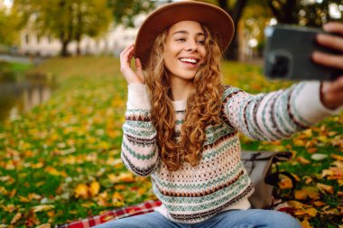 Şapkalı neşeli kadın, sonbahar parkında kırmızı bir battaniyenin üzerinde otururken telefonuyla selfie çekiyor. Elinde bir buket parlak yaprak bloguyla doğayı seven genç bir kadın. Hafta sonu konsepti.