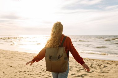 Evrak çantalı tatlı bir kadın deniz kıyısında ya da okyanusta duruyor ve günbatımının tadını çıkarıyor. Gezgin açık havada özgürlüğü hissediyor. Macera, doğa konsepti. Yaşam biçimi.