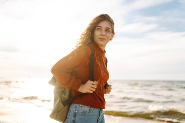 Evrak çantalı tatlı bir kadın deniz kıyısında ya da okyanusta duruyor ve günbatımının tadını çıkarıyor. Gezgin açık havada özgürlüğü hissediyor. Macera, doğa konsepti. Yaşam biçimi.