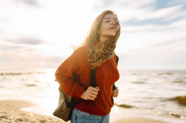 Evrak çantalı tatlı bir kadın deniz kıyısında ya da okyanusta duruyor ve günbatımının tadını çıkarıyor. Gezgin açık havada özgürlüğü hissediyor. Macera, doğa konsepti. Yaşam biçimi.
