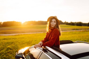 Şapkalı kadın gezgin, arabasının camından sarkıyor ve huzurlu kırsal manzaranın arka planında günbatımının tadını çıkarıyor. Araba macerasında güzel bir kadın dışarıda olmanın özgürlüğünü hisseder. Etkin yaşam biçimi.