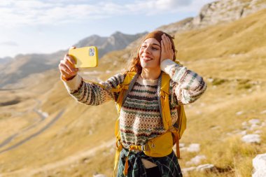 Renkli sırt çantalı kadın yürüyüşçü, resim gibi dağ manzarasının arka planına selfie çeker. Yürüyüş yapan kadın bloglarında günbatımı macerasının tadını çıkarıyor. Bloglama, doğa kavramı.