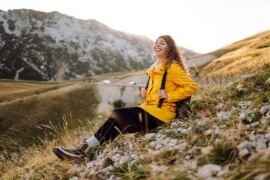 Parlak yağmurluklu neşeli kadın gün batımında dağlık araziyi keşfediyor. Sırt çantalı genç bayan gezgin özgürlüğü hissediyor ve dağ manzarasının tadını çıkarıyor. Doğa, macera konsepti.