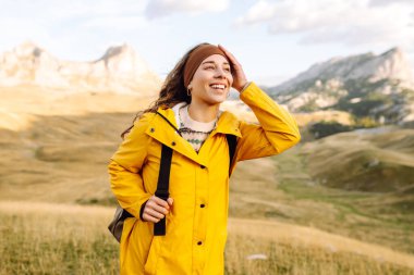 Parlak yağmurluklu neşeli kadın gün batımında dağlık araziyi keşfediyor. Sırt çantalı genç bayan gezgin özgürlüğü hissediyor ve dağ manzarasının tadını çıkarıyor. Doğa, macera konsepti.