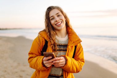 Kumsalda sarı ceketli genç bir kadın ve plajda günbatımının tadını çıkarıyor. Neşeli bir kadın telefonda mesaj atıyor, sahilde yürüyor. Seyahat, teknoloji konsepti.
