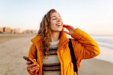 Kumsalda sarı ceketli genç bir kadın ve plajda günbatımının tadını çıkarıyor. Neşeli bir kadın telefonda mesaj atıyor, sahilde yürüyor. Seyahat, teknoloji konsepti.