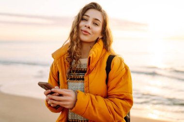 Kumsalda sarı ceketli genç bir kadın ve plajda günbatımının tadını çıkarıyor. Neşeli bir kadın telefonda mesaj atıyor, sahilde yürüyor. Seyahat, teknoloji konsepti.