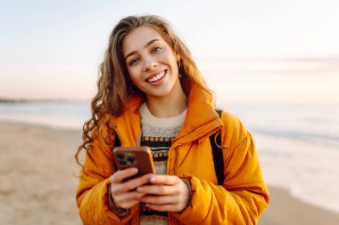 Kumsalda sarı ceketli genç bir kadın ve plajda günbatımının tadını çıkarıyor. Neşeli bir kadın telefonda mesaj atıyor, sahilde yürüyor. Seyahat, teknoloji konsepti.