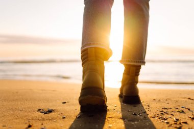 Kumlu bir gün batımı plajında yürüyüş botlarıyla dikilirken. Güneş ışığında botları olan ve deniz manzarasının tadını çıkaran bir kadın gezgin. Macera ve doğa kavramı.