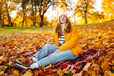 Parlak ceketli mutlu bir kadın, sonbahar yaprakları arasında bir battaniyenin üzerinde otururken gün batımının tadını çıkarıyor. Sonbahar parkında battaniyeye sarılmış bir gezgin. Doğa, keyif ve rahatlama kavramı..