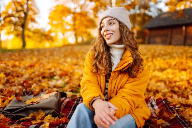 Parlak ceketli mutlu bir kadın, sonbahar yaprakları arasında bir battaniyenin üzerinde otururken gün batımının tadını çıkarıyor. Sonbahar parkında battaniyeye sarılmış bir gezgin. Doğa, keyif ve rahatlama kavramı..