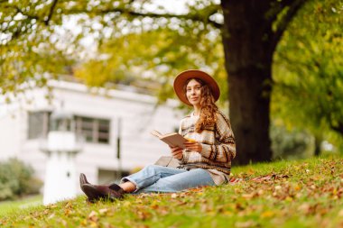 Nehir kenarındaki bir açıklıkta genç bir kadın sonbahar parkında rahat bir battaniyenin üzerinde otururken kitap okuyor. Şapkalı güzel bir kadın sonbaharı ve açık havada kitap okumayı sever. Doğa, rahatlama ve hafta sonu konsepti.