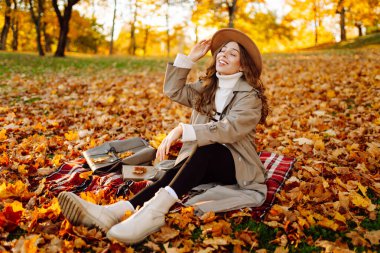 Yağmurluk ve şapkalı mutlu kadın gün batımında kırmızı battaniyeye oturmuş sonbahar parkının keyfini çıkarıyor. Sırt çantalı, açık havada dinlenen güzel bir bayan gezgin. Doğa konsepti, hafta sonu.