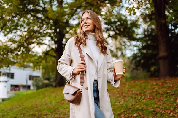 Paltolu genç bir kadın güneşli sonbahar parkında elinde bir fincan kahveyle yürüyor. Günbatımının ve açık havada sıcak içeceğin tadını çıkaran güzel bir turist. Doğa, güzellik, içki konsepti.