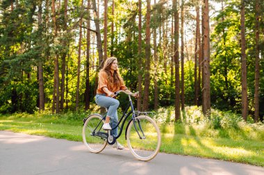 Yoğun güneşli bir ormanda bisiklet süren genç bir kadın. Doğanın tadını çıkaran ve gün batımında bisiklet süren mutlu bir kadın. Hafta sonu konsepti. Etkin yaşam biçimi.