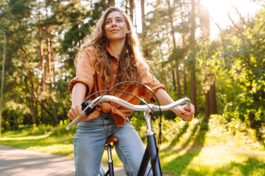 Yoğun güneşli bir ormanda bisiklet süren genç bir kadın. Doğanın tadını çıkaran ve gün batımında bisiklet süren mutlu bir kadın. Hafta sonu konsepti. Etkin yaşam biçimi.