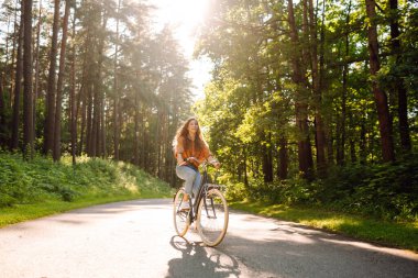 Yoğun güneşli bir ormanda bisiklet süren genç bir kadın. Doğanın tadını çıkaran ve gün batımında bisiklet süren mutlu bir kadın. Hafta sonu konsepti. Etkin yaşam biçimi.