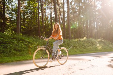 Yoğun güneşli bir ormanda bisiklet süren genç bir kadın. Doğanın tadını çıkaran ve gün batımında bisiklet süren mutlu bir kadın. Hafta sonu konsepti. Etkin yaşam biçimi.